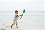 Young boy standing on beach, holding up shovel, looking down