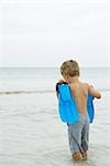 Young boy standing knee deep in water, carrying flippers, rear view