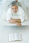 Mature man lying on stomach on chaise longue in front of book, looking up at camera