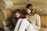 Teen girl and preteen boy sitting side by side, reading and playing video game