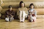 Drei junge Geschwister sitzen nebeneinander, lesen und spielen Videospiel