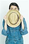 Teenage girl holding straw hat in front of face, looking away