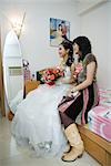 Bride sitting on bed with friend, looking away, smiling, full length