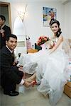 Groom putting shoe on bride's foot in bedroom, both smiling at camera, full length