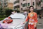 Bride dressed in traditional Chinese clothing, standing next to decorated car, smiling at camera