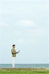 Man standing on low wall using laptop, body of water in background, full length