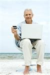 Senior man using laptop on beach, making on-line purchase with credit card