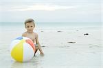 Little boy crouching behind ball at the beach, smiling at camera