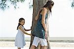 Two sisters standing beside tree trunk, one holding on to the other's shirt with eyes closed