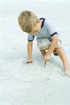 Petit garçon accroupi sur la plage, de dessin dans le sable avec bâton