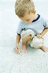 Little boy crouching at the beach, drawing in sand with stick