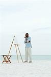 Senior man standing in front of blank canvas at the beach, holding paint brushes