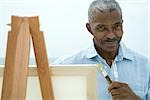 Senior man with hand under chin, holding paint brushes, smiling at camera