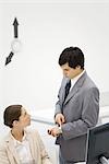 Male supervisor standing beside female employee, pointing at wristwatch, clock hands on wall in background