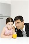 Father and daughter sharing glass of orange juice, drinking from straws, looking at camera