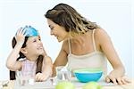 Mother and daughter sitting at table, girl placing bowl on her head, both smiling