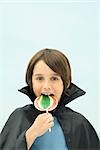 Boy wearing vampire cape, eating large lollipop, portrait