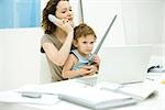 Young mother sitting at desk using phone while son sits on lap, holding sword