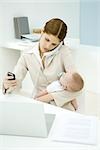 Young working mother holding baby, talking on landline phone and looking at cell phone