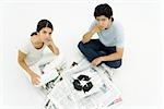 Young couple sitting beside stack of newspapers stenciled with recycling symbol, looking up at camera