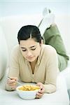 Young woman lying on stomach on couch, holding cereal bowl, looking down