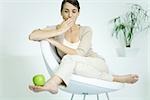 Young woman slouching in chair, balancing apple on foot, covering mouth