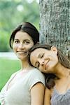 Mother and teenage daughter leaning against tree, girl's head on mother's shoulder, smiling