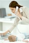 Professional woman using laptop computer and phone, infant lying on desk