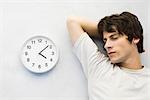 Young man leaning against wall beside clock, eyes closed