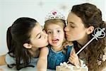 Little girl dressed as princess, being kissed on cheeks by mother and sister