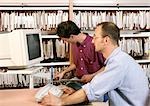 Man using computer, other man looking at files on shelves