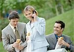 Three business people holding sandwiches outdoors, one using cell phone