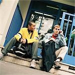 Young men sitting on steps with backpacks, one using cell phone.
