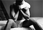 Man sitting on bed, t-shirt around neck, hand to face, black and white.