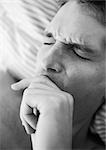 Man's face with eyes closed and furrowed brow, hand over mouth, lying on bed, close-up, black and white.