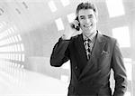 Businessman using cellular phone in corridor, smiling at camera, black and white