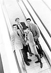 Group of business people on moving walkway smiling at camera, business woman in front on cell phone, b&w.