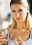 Woman holding pill and glass, looking at camera.