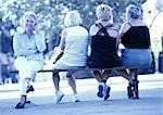 Four women sitting on a bench, one using phone, three seen from the back