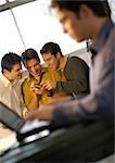 Three men looking at hand held computer, fourth man using laptop in foreground