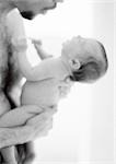 Father holding infant against bare chest, side view, b&w