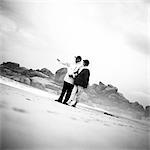 Mature couple standing on beach, man pointing, b&w