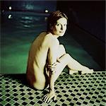 Nude woman sitting by edge of swimming pool, rear view