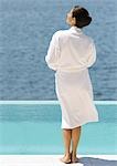 Woman in bathrobe standing near pool overlooking sea, rear view