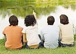 Four friends sitting near the edge of a pond, one pointing, rear view