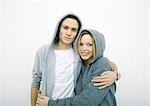 Young couple standing with arms around each other, wearing hooded sweatshirts, looking at camera