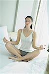 Woman sitting in lotus position on top of bed, eyes closed