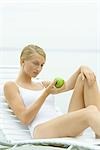 Teenage girl in swimsuit sitting on lounge chair, holding apple