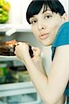 Young woman taking slice of cake from refrigerator, looking over shoulder at camera