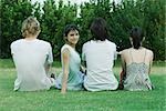 Group of young friends sitting on grass, rear view, one young woman looking over shoulder at camera